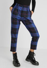 Pantalons en tartan bleu et noir avec des accents rouges, taille haute, présentant une coupe ajustée et des plis, associés à des bottes de combat brillantes noires.
