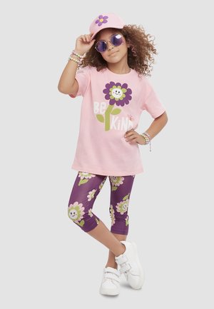 Dziewczyna o kręconych włosach nosi różową czapkę z daszkiem i okulary przeciwsłoneczne, różowy t-shirt z napisem "Be Kind" i uśmiechniętym kwiatem, fioletowe legginsy w kwiaty, białe trampki i bransoletki.
