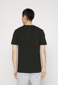 En man sedd bakifrån som bär en enkel svart t-shirt med kort ärm och ljusgråa sweatpants mot en helt vit bakgrund.