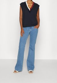 Top senza maniche blu navy con strisce bianche e bordeaux a contrasto sul colletto, abbinato a jeans chiari a zampa d'elefante e sandali bianchi.