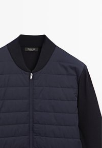 Navy quilted bomber jacket with a ribbed collar, featuring a silver zipper, and contrasting smooth fabric on the sleeves.