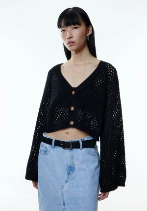Zwarte opengehaakte cropped cardigan met lange mouwen, voorzien van een V-hals en drie knopen aan de voorkant, gecombineerd met een lichtblauwe denim rok.