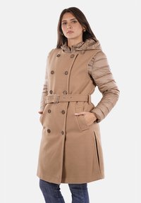 Cappotto beige doppiopetto con cintura, dotato di cappuccio e maniche trapuntate. Tessuto liscio, tasche laterali e bottoni marroni.