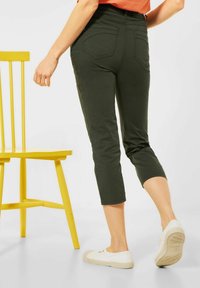 Person som bär korta mörkgröna byxor och vita sneakers står vid en ljusgul trästol mot en vit bakgrund.