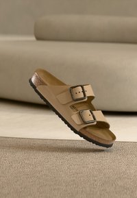 Μπεζ σανδάλι με δύο λουράκια, μαύρα αγκράφες και φελλό στην σόλα, αιωρούμενο πάνω από υφασμένο χαλί σε καθιστικό με ουδέτερους τόνους.