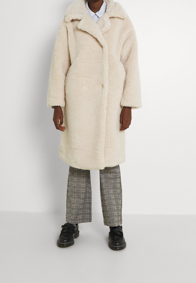 Abrigo de forro polar color crema con un gran cuello de pico, botones de doble botonadura y hombros caídos, combinado con pantalones de cuadros y zapatos negros.