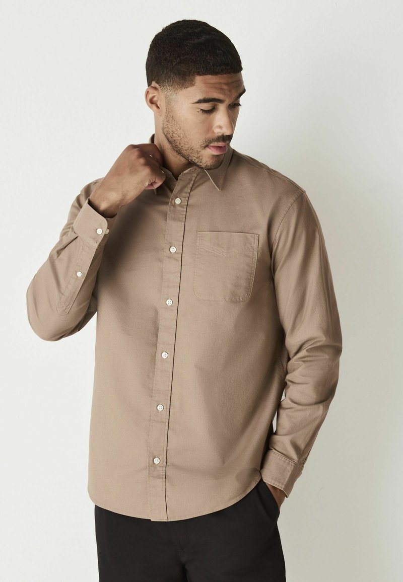 Camisa de botones marrón con mangas largas, un bolsillo en el pecho y botones blancos. Textura suave y corte relajado, llevada con pantalones negros.