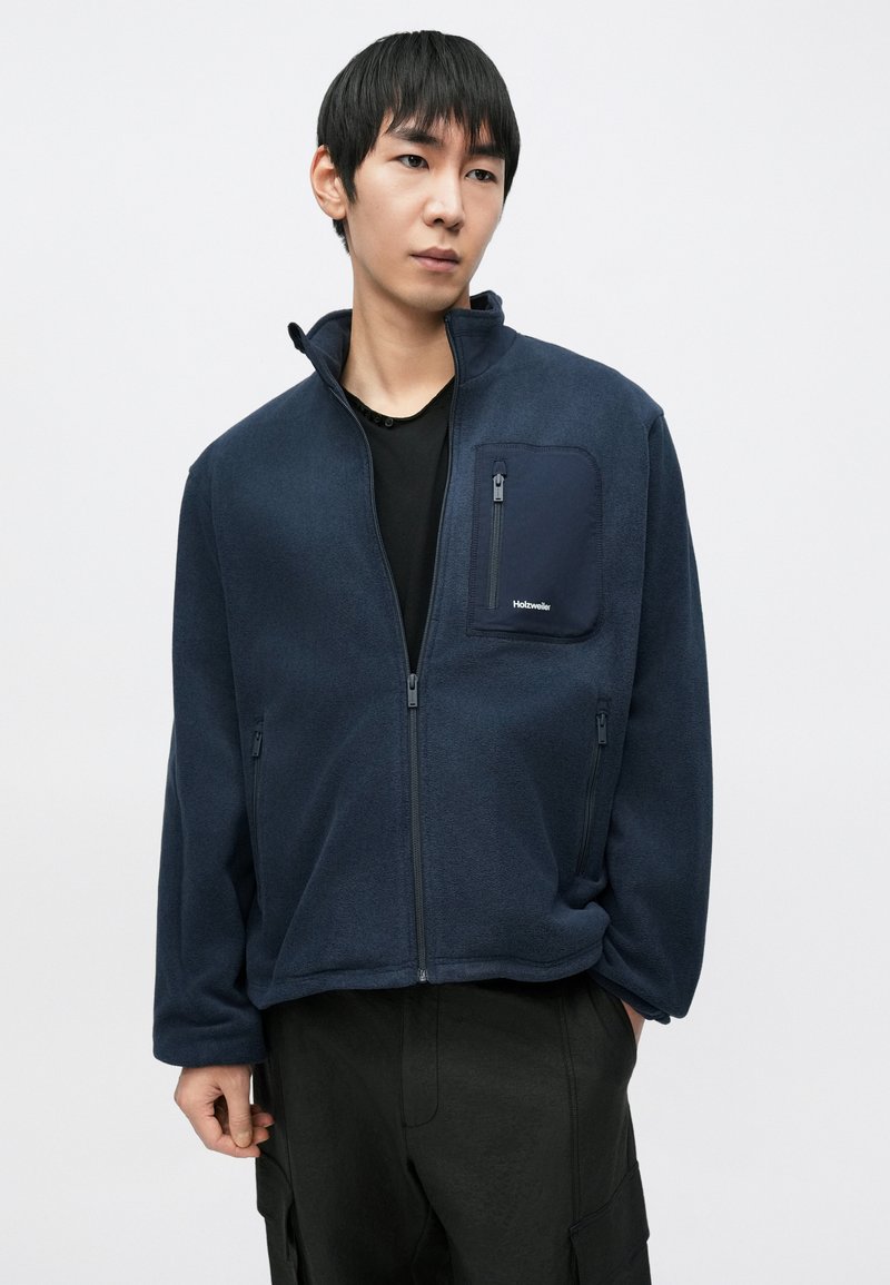 Veste polaire bleue marine avec fermeture éclair et poche poitrine. Caractérisée par un col montant et des poches latérales zippées. Texture lisse et coupe décontractée.