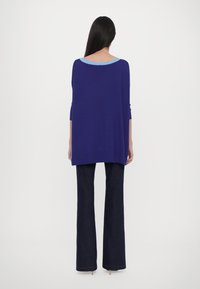 Femme aux longs cheveux foncés portant un haut bleu ample avec un col bleu clair et un pantalon large foncé, debout de dos sur fond blanc.