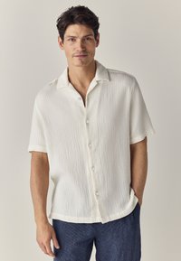 Camicia a maniche corte, con bottoni, in morbido tessuto bianco sporco, caratterizzata da un colletto aperto e una vestibilità rilassata. Abbinata a pantaloni in denim.