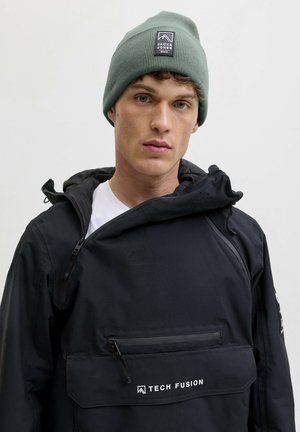 Jonge man met een groene beanie met het label "Jack & Jones" en een zwart Tech Fusion-jack, staand tegen een effen lichte achtergrond.