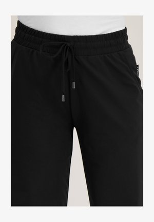 Zwarte broek met trekkoord en elastische tailleband, metalen uiteinden aan de koorden en zijkanten met zakken, gedragen met een wit bovenstuk.