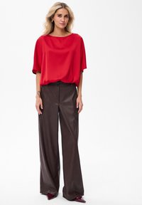 Vrouw die staat in een losse rode top, high-waisted broek van bruin leer met wijd uitlopende pijpen, en puntige donkerrode schoenen op een witte achtergrond.