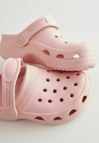 Las zuecos de plástico rosa cuentan con una punta cerrada, una correa trasera ajustable, agujeros de ventilación y un detalle texturizado a lo largo de los lados y la base.