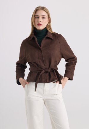 Donna che indossa una giacca marrone con cintura sopra un dolcevita verde scuro e pantaloni bianchi, in piedi con le mani nelle tasche contro uno sfondo semplice.