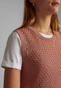 Rosa stickad väst med upphöjd prickmönster, rund halsringning, ärmlös design, lagd över en vit skjorta med uppvikta ärmar.