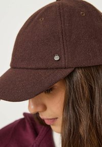 Casquette en laine brune avec un bord incurvé, dotée d'un petit logo métallique en accent. La vue en gros plan met en évidence la texture et les détails de couture.