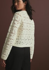 Haaktrui in crème met opengewerkte bloemenpatronen, lange mouwen en een cropped zoom, gecombineerd met zwarte broek tegen een neutrale achtergrond.