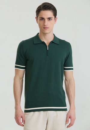 Mann trägt ein dunkelgrünes kurzärmeliges Poloshirt mit weißen Streifen an Ärmeln und Saum, steht vor einem einfarbigen hellen Hintergrund.