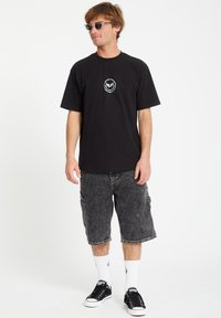 Czarny bawełniany t-shirt z centralnym graficznym wzorem w kształcie okręgu, zestawiony z ciemnoszarymi szortami cargo. Noszony z czarnymi butami sportowymi i białymi skarpetkami.