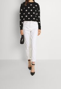 Svart tröja med ljusa prickar, vita högmidjade jeans och svarta pumps med spetsig tå. Modellen håller en liten texturerad svart handväska.