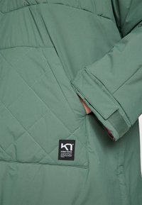 Chaqueta acolchada verde con un acabado texturado, que cuenta con un bolsillo de parche y una etiqueta de marca negra en la parte inferior derecha del diseño.