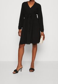 Vestido negro con escote en V, mangas largas y detalle fruncido en la cintura. Tela lisa, largo hasta la rodilla, combinado con sandalias de tacón trenzadas y un bolso de mano.