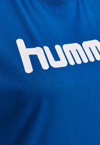 Blå bomullströja med en stor vit "humm"-logotyp. Tyget är slätt, med rund hals och korta ärmar.