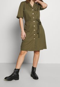 Robe chemise vert olive à manches courtes, boutons devant, deux poches poitrine et ceinture à la taille. Bottines noires avec un détail zippé.
