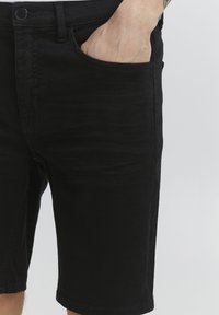 Shorts negri din denim cu un design cu cinci buzunare, ușor elastici și o croială dreaptă cu un tiv brut. Descrierea este un prim-plan al buzunarului din față.
