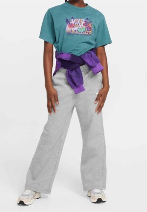 Personne portant un t-shirt graphique bleu sarcelle, un sweat-shirt violet attaché à la taille, un pantalon de survêtement gris à larges jambes et des baskets blanches, debout devant un fond blanc.