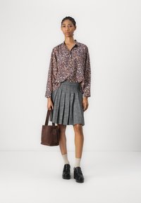 Leopardmønstrete skjorte med knappestolpe, grå plissert skjørt, brun tote bag, svarte kraftige sko, kremfargede strømper, modell står mot en hvit bakgrunn.
