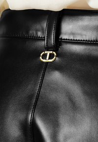 Schwarze Lederhosen mit glänzender Textur, versehen mit einer goldenen ovalen Schnalle mit der Aufschrift "TWINSSET" und sichtbaren Nahtdetails am Bund.