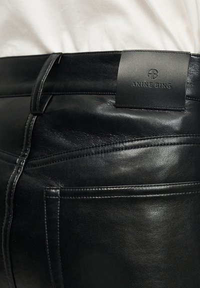 Close-up de calças de couro pretas com bolso traseiro e etiqueta de couro da marca Anine Bing em relevo na cintura.