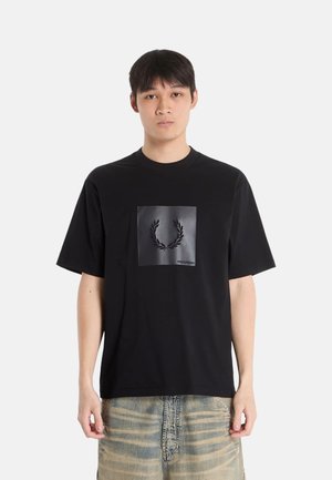 Joven de pie vistiendo una camiseta negra Fred Perry con un logo de corona de laurel y vaqueros beige a rayas sobre un fondo blanco liso.