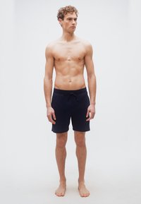 Νεαρός άνδρας με σγουρά μαλλιά, όρθιος ξυπόλητος, φοράει σκούρα μπλε ναυτικά σορτς με κορδόνι, πάνω σε απλό φωτεινό φόντο.