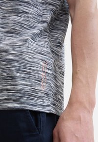 Gray and white striped short-sleeve shirt with a pink logo, featuring a soft texture and a relaxed fit, shown alongside a hand in navy fabric.