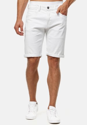 Homme portant un short en denim blanc retroussé et des baskets blanches, debout avec une main dans la poche, devant un fond uni.