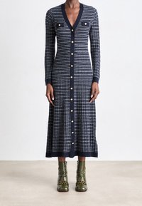 Pletené šaty s vertikálními námořnickými a bílými pruhy, výstřihem do V, dlouhými rukávy, zlatými knoflíkovými detaily a dvěma náprsními kapsami. V páru s zelenými kotníkovými botami.