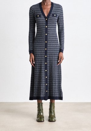 Strickkleid mit vertikalen navyblauen und weißen Streifen, V-Ausschnitt, langen Ärmeln, goldfarbenen Knopfakzenten und zwei Brusttaschen. Kombiniert mit grünen Stiefeletten.
