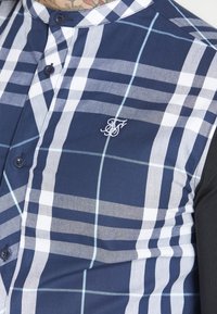 Short-sleeve shirt featuring a navy and white plaid pattern, with embroidered logo, featuring a band collar and contrasting black sleeves.