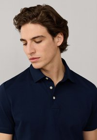Joven con cabello castaño ondulado que lleva una camisa tipo polo azul marino oscuro, mirando hacia abajo contra un fondo liso gris claro.
