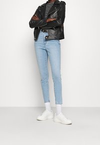 Person som bär ljusblå skinny jeans, svart skinnjacka, randig tröja, vita strumpor och vita sneakers, står med armarna i kors.