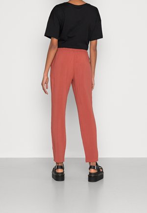 Person trägt rostfarbene Hose, schwarzes kurzärmeliges Hemd und schwarze Plateausandalen, steht vor einem hellgrauen Hintergrund.