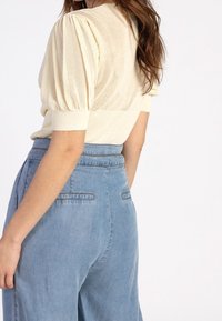 Krémový síťovaný top s krátkým rukávem a nařasenými rukávy, spárovaný se světle modrými džínovými kalhotami s vysokým pasem, které mají zadní kapsy a volný střih.