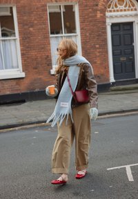 Veste en cuir marron, écharpe bleu clair à franges, pantalon large beige, ballerines rouges, tenant une tasse orange dans un décor de rue en ville.
