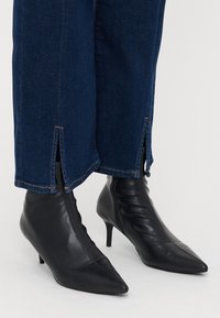 Botas de tornozelo em couro preto com bico fino e salto fino, combinadas com jeans escuros com detalhe de bainha desfiada.