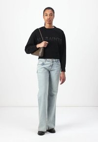 Felpa nera cropped con lettering testurizzato "ALLSAINTS", abbinata a jeans a gamba larga di colore blu chiaro e scarpe nere; tiene in mano una borsa marrone.