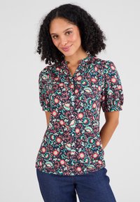 Blusa a bottoni floreale con maniche a palloncino corte, con uno sfondo blu navy, presentante motivi floreali colorati in rosa, verde e crema su un tessuto leggero.