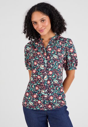Blouse à manches courtes bouffantes et à boutons floraux, avec un fond marine, des motifs floraux colorés en rose, vert et crème sur un tissu léger.